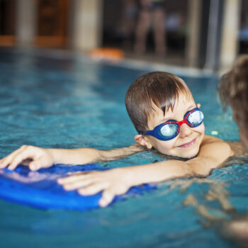 Happy little boy during swimming lesson smiles at his mother. The boy is aged 6 and is wearing swimming goggles. The boy is using a kickboard.